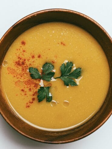 A creamy yellow lentil soup garnished with fresh parsley and sprinkled with red chili flakes, served in a rustic brown bowl against a white background.