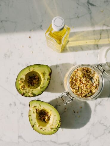 A top-down view of two halved avocados drizzled with oil and sprinkled with mixed seeds, next to a small bottle of golden oil and a glass jar filled with assorted seeds, all placed on a white marble surface with soft shadows.