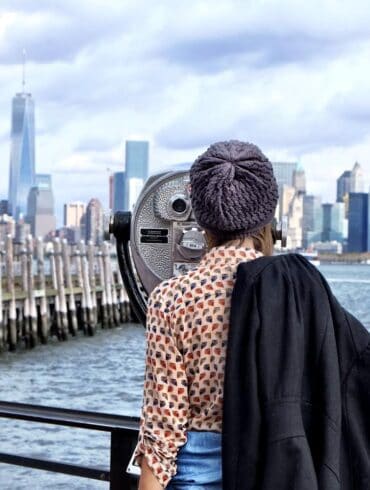 Health coach and wellness blogger valerie alvarez with a knitted hat looking through a coin-operated binoculars towards the New York City skyline, with the One World Trade Center prominently in view, against a backdrop of a cloudy sky.
