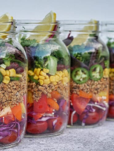 Colorful meal prepped mason jars filled with layered fresh ingredinets of cabbage, bell pepper, quinoa, corn, jalapenos,black beans, cilantro, lettuce and lime.