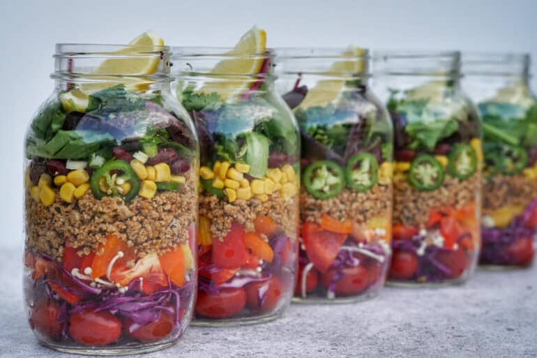 Colorful meal prepped mason jars filled with layered fresh ingredinets of cabbage, bell pepper, quinoa, corn, jalapenos,black beans, cilantro, lettuce and lime.