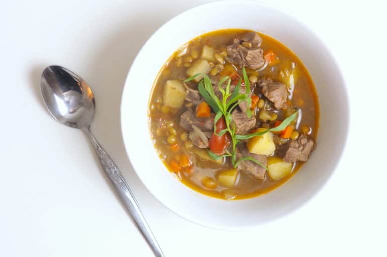Overhand shot of a bowl of lamb lentil stew with potatoes, tomatoes and sprig of fresh tarragon and a silver spoon to the left on top of a white surface.