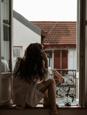 A woman sitting thoughtfully on a windowsill, wearing a white shirt, with her back to the camera, looking out over a European-style neighborhood. She has one hand to her head, suggesting contemplation or relaxation, beside her a small French press with coffee on the windowsill.
