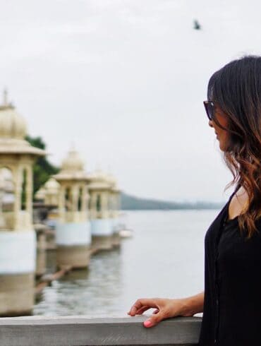 A woman in a casual black top and sunglasses stands by a lakeside railing, gazing out at an ornate building with traditional domes and a serene water backdrop in Udaipur, India, reflecting a moment of peaceful contemplation.