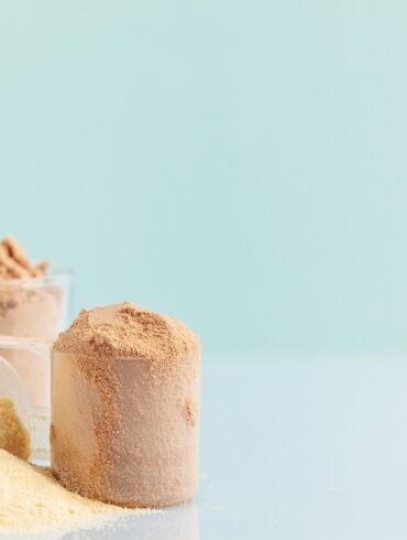 A variety of protein powders displayed against a pastel blue background. On the left is a translucent scoop filled with a pale yellow powder, likely representing a vanilla flavor. In the middle, there is a scoop with a darker, chocolate-colored powder, and on the right, a scoop overflows with a beige powder, possibly indicating an alternative flavor or ingredient intended to be sugar-free.