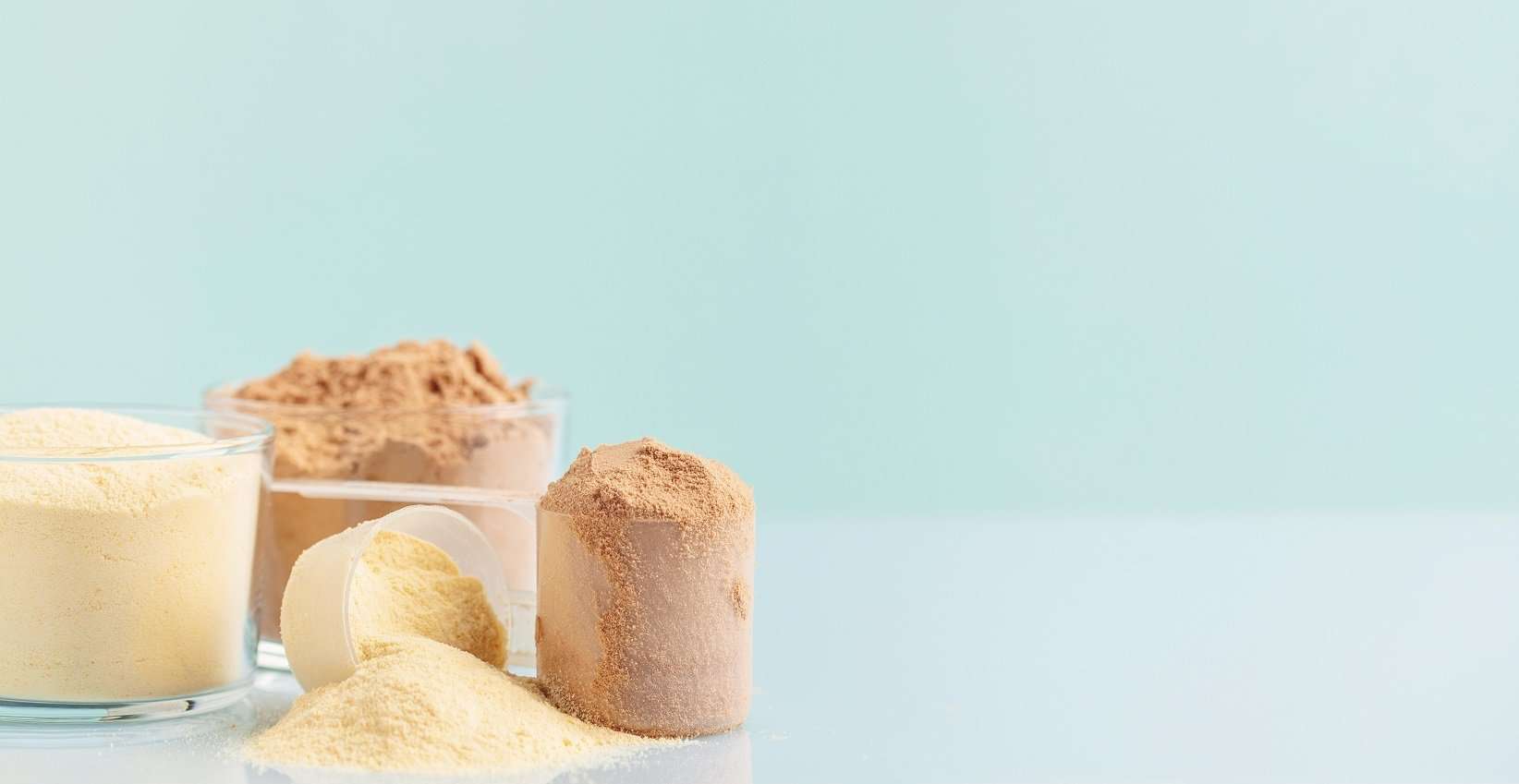 A variety of protein powders displayed against a pastel blue background. On the left is a translucent scoop filled with a pale yellow powder, likely representing a vanilla flavor. In the middle, there is a scoop with a darker, chocolate-colored powder, and on the right, a scoop overflows with a beige powder, possibly indicating an alternative flavor or ingredient intended to be sugar-free.
