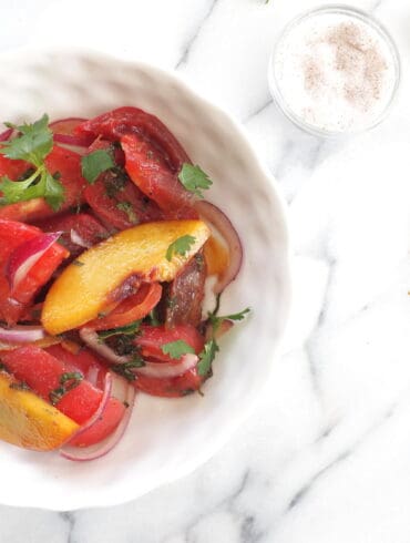 A close-up image of a vibrant Heirloom Tomato and Peach Salad in a white bowl, showcasing slices of red and green-striped tomatoes, golden peaches, and red onions, garnished with fresh green cilantro leaves on a marble surface.