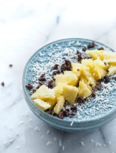 A bowl of Blue Majik chia pudding. The pudding has a distinct blue color, indicative of the blue-green algae used in its creation. It's topped with bright yellow pineapple chunks that add a pop of color, and scattered cacao nibs that provide a rich, dark contrast to the light blue background of the pudding. The bowl sits on a marble surface, suggesting a modern, health-conscious lifestyle