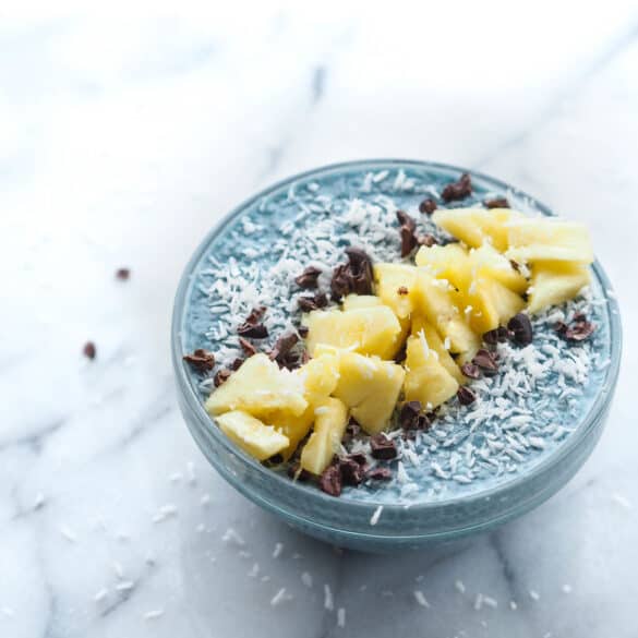 A bowl of Blue Majik chia pudding. The pudding has a distinct blue color, indicative of the blue-green algae used in its creation. It's topped with bright yellow pineapple chunks that add a pop of color, and scattered cacao nibs that provide a rich, dark contrast to the light blue background of the pudding. The bowl sits on a marble surface, suggesting a modern, health-conscious lifestyle
