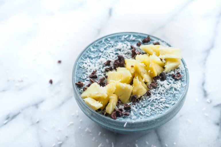A bowl of Blue Majik chia pudding. The pudding has a distinct blue color, indicative of the blue-green algae used in its creation. It's topped with bright yellow pineapple chunks that add a pop of color, and scattered cacao nibs that provide a rich, dark contrast to the light blue background of the pudding. The bowl sits on a marble surface, suggesting a modern, health-conscious lifestyle